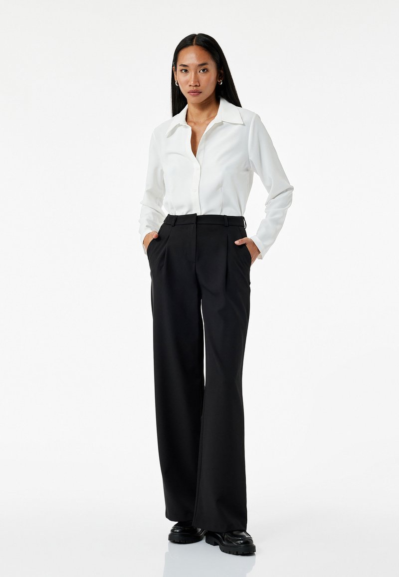 Witte blouse met lange mouwen en knopen, gecombineerd met hoge taille zwarte wijde broek. De outfit heeft een gestroomlijnd ontwerp en een zachte textuur.