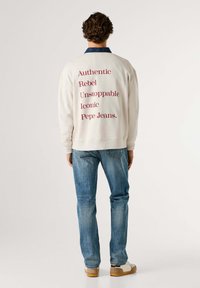 Felpa bianca con scritta rossa: "Authentic Rebel Unstoppable Iconic Pepe Jeans." Abbinata a jeans blu e scarpe beige.