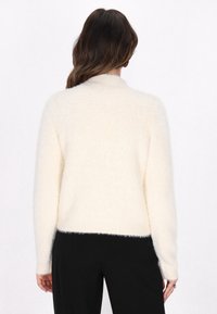 Crème-kleurige, fluffy trui met een hoge hals en lange mouwen, voorzien van geribbelde manchetten en zoom, gecombineerd met zwarte broek.