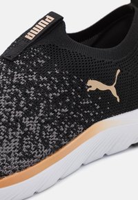 Baskets de sport noires avec une tige en maille texturée, dotées d'un logo Puma doré et d'une semelle blanche avec une touche de brun. Vue en gros plan.