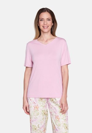 Lächelnde Frau, die ein hellrosa Kurzarm-V-Ausschnitt-Shirt und eine Hose mit floralem Muster trägt, vor einem einfarbigen hellen Hintergrund steht.