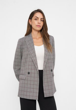 Mujer con cabello largo y castaño, con pendientes de aro dorados, blazer gris a cuadros de doble botonadura, top blanco y pantalones negros, posando frente a un fondo blanco.