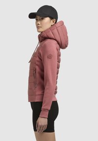 Roze hoodie met een puffy gewatteerde capuchon, ritszak aan de zijkant en geribde manchetten. Gecombineerd met een zwarte pet en zwarte shorts.
