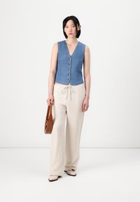Denimvest med V-hals og fem knappelukkinger, kombinert med lysebeige vide bukser og en brun skulderveske. Glatte teksturer gjennomgående.