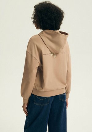 Osoba o kręconych włosach ubrana w beżową bluzę z napisem "Mediterranean Wildlife" na plecach, w ciemnoniebieskich dżinsach, zwrócona tyłem.