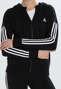 Sweat à capuche noir zippé en tissu doux, avec des rayures blanches sur les manches et un petit logo en haut à gauche. Capuche avec cordon de serrage incluse.