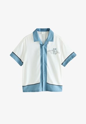 Dreng iført en hvid og blå skjorte og shorts går på en stenfod ved siden af en hvid væg i klart sollys.