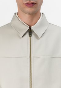Lichtbeige ritsjack met een verhoogde kraag, voorzien van een gouden rits en nauwkeurige stikseldetails. Vlotte textuur, minimalistisch ontwerp.