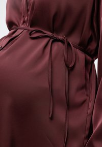 Robe en satin rouge foncé avec un petit nœud à la taille, des détails de boutons et des manches longues en gros plan.