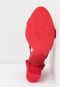 Chaussure à talons hauts rouge avec des initiales en écriture cursive en relief sur la semelle, positionnée semelle vers le haut contre un mur d'angle blanc.