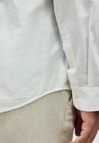 Κοντινό πλάνο στο χέρι ενός ατόμου δίπλα στο σώμα, που φορά λευκό πουκάμισο με μακριά μανίκια και ανοιχτό μπεζ υφασμάτινο παντελόνι.