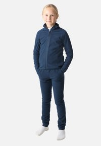 Laivansinen fleece-harjoitusasu, jossa on korkea kaulus, vetoketjukiinnitys ja joustavat hihansuut. Housuissa on tiukka istuvuus ja ne on suunniteltu samansävyiseksi.