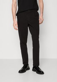 Černé bavlněné kalhoty s kapsami, rovný střih a minimální švy. V kombinaci s lesklými černými botami, na jednolitém pozadí.