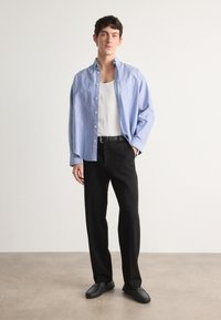 Chemise à rayures bleues et blanches, coupe décontractée, associée à un pantalon noir et des chaussures noires à enfiler. Débardeur blanc en dessous.