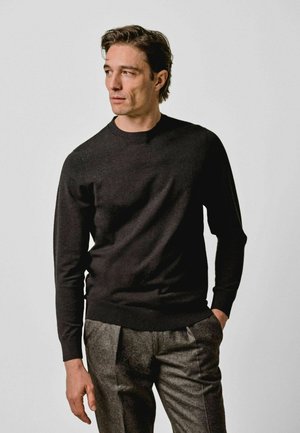 Uomo che indossa una camicia scura a maniche lunghe e pantaloni grigi, in piedi con una mano in tasca, che guarda di lato davanti a uno sfondo semplice.