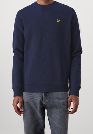 Mann trägt ein marineblaues Sweatshirt mit langen Ärmeln und einem kleinen gelben Vogel-Logo auf der Brust sowie ausgewaschene blaue Jeans, steht vor einem hellen Hintergrund.