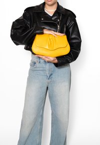 Geel leren clutch met een gestructureerd oppervlak, met een verzamelde bovenkant en een minimalistisch ontwerp. Model houdt het tegen een zwarte jas.