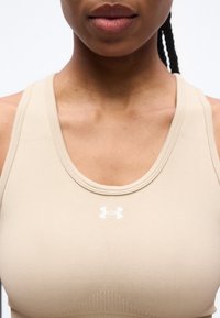 Γυναίκα που φοράει μπεζ αθλητικό σουτιέν Under Armour, εμφανιζόμενη από τους ώμους μέχρι τα χείλη, με πλεξούδα εμφανή πάνω στον ώμο.