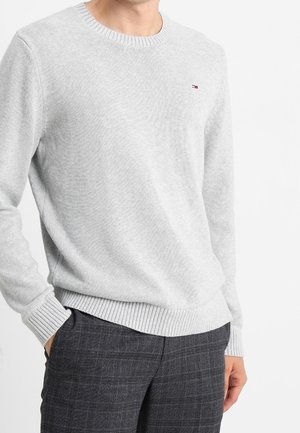 Hellgrauer Strickpullover mit Rundhalsausschnitt, gerippten Bündchen und Saum, versehen mit einem kleinen Logo auf der Brust, kombiniert mit karierten Hosen.