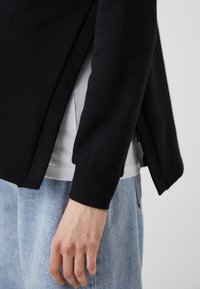 Black sweatshirt with a relaxed fit, ribbed cuffs, and side slits, layered over a white shirt, paired with light blue denim shorts.