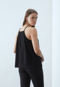 Svart plissert tank top med tynne stropper, har en løs passform og rett søm. Båret med svarte bukser. Bakgrunnen er en lysekket vegg.