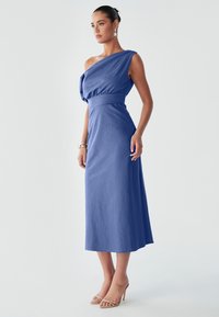 Modelka v modrých šatech na jedno rameno stojí elegantně, na nohách má holenové sandály, s neutrálním pozadím, které zdůrazňuje její stylovou pózu.
