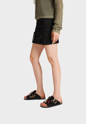 Donna con minigonna nera arricciata, maglione verde oliva in rete e sandali neri slip-on con perline colorate e fibbia su sfondo chiaro.