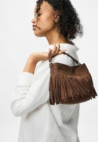 Brūnas zamšas rokassoma ar frīzu detaļām un īsu rokturi, kuru tur cilvēks, kas valkā baltu kreklu. Gluda tekstūra, noapaļota forma.