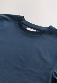 Tamnoplava majica s okruglim izrezom i vidljivom bijelom etiketom unutar ovratnika na svijetloj pozadini.