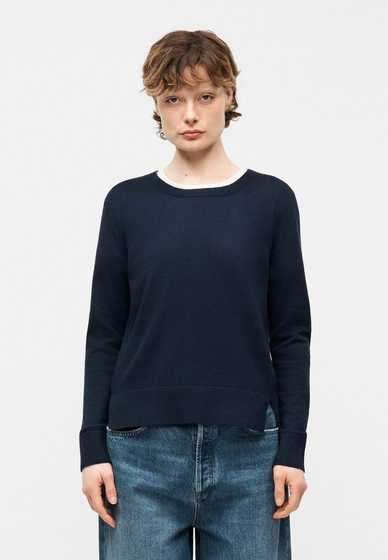 Jeune personne aux cheveux courts portant un pull à manches longues bleu marine par-dessus une chemise blanche et un jean large bleu, debout devant un fond uni.