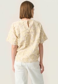Texturierte cremefarbene Bluse mit floraler Stickerei, kurzen Ärmeln, runder Ausschnitt und kleinem Schlüssellochverschluss hinten. Kombiniert mit weißen Jeans.
