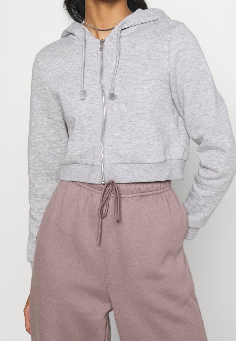 Sudadera gris de corte corto hecha de tela suave, con una cremallera frontal y cordones en la capucha. Combinada con pantalones de chándal rosas con cordones.