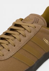 Καφέ πάνινο παπούτσι από σουέντ και υφασμάτινο με έξι κορδόνια, τρεις λωρίδες στο πλάι και λογότυπο «Gazelle» σε χρυσό. Λαστιχένια σόλα με σκούρα έντονη λεπτομέρεια.