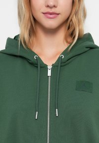 Zelená mikina na zips s rebrovanou textúrou, striebornými doplnkami a čiernou logom na oblasti hrude. Obsahuje nastaviteľné šnúrky.