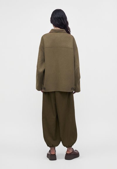 Mujer vista desde atrás, con una chaqueta verde oliva de gran tamaño y pantalones anchos a juego con puños fruncidos, de pie sobre un fondo blanco liso.
