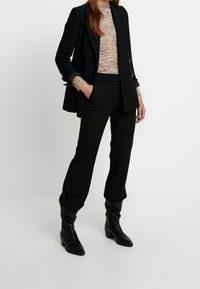 Blazer negro a medida sobre un suéter texturizado multicolor, combinado con pantalones negros de talle alto y botas negras hasta la rodilla.