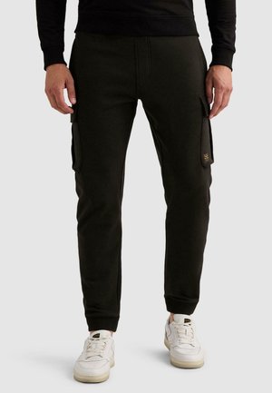 Homme portant un pantalon cargo jogger noir avec des poches latérales et des baskets blanches, debout devant un fond clair uni.