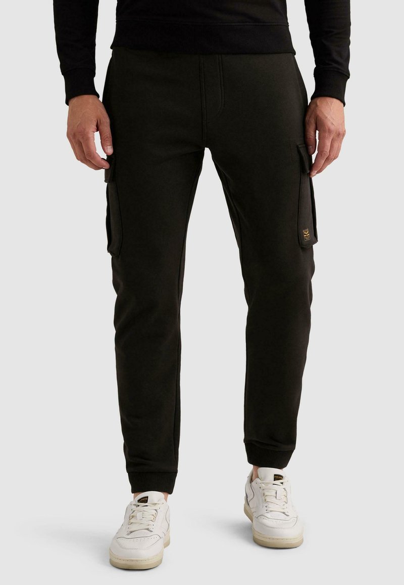 Άνδρας που φοράει μαύρο cargo παντελόνι jogger με πλαϊνές τσέπες και λευκά αθλητικά, στέκεται μπροστά σε απλό φωτεινό φόντο.