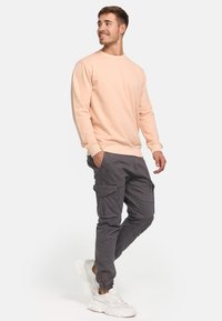 Lys ferskenfarvet sweatshirt med lange ærmer, parret med mørkegrå cargo-bukser med sidelommer. Hvide sneakers med teksturerede detaljer.