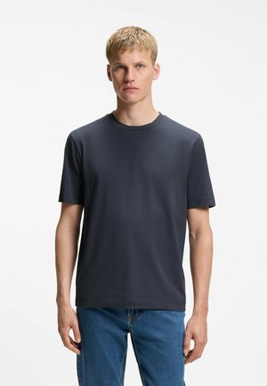 Ung mand med kort blondt hår står forlæns, iført en ensfarvet mørkeblå t-shirt og blå jeans mod en hvid baggrund.