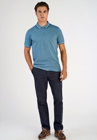 Niebieska koszulka polo z zamkiem błyskawicznym i białymi wykończeniami, w połączeniu z granatowymi spodniami i brązowymi butami typu boat. Gładka tekstura, casualowy design.