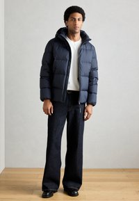 Casaco acolchoado azul-marinho com capuz, apresentando textura acolchoada e fecho de correr frontal, usado sobre uma camisa branca e calças de ganga escuras.