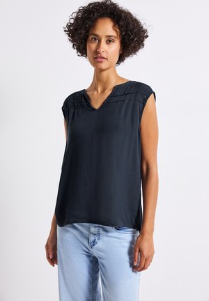 Top sans manches bleu marine avec un tissu texturé, présentant un décolleté en V et des coutures décoratives le long des épaules. Assorti à un jean bleu clair.