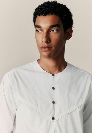 Jaunas vyras su garbanotais juodais plaukais, vilkintis baltą marškinių su sagomis, žiūrintis į šoną priešais paprastą smėlio spalvos foną.