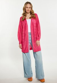 Rožinė megzta kardiganas su atviru priekiu ir išskirtiniais raštais, derinama su šviesiai mėlynomis plačiomis kelnėmis ir oranžiniais platforminiais sandalais.