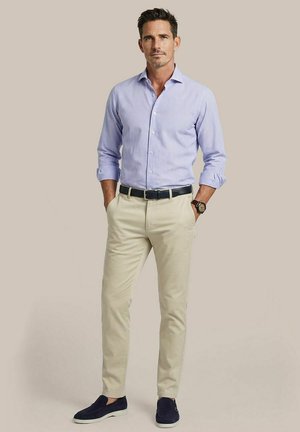 Man draagt lichtblauw gestreept overhemd, beige broek, zwarte riem, zwart horloge en donkerblauwe instapschoenen, staat met handen in de zakken.