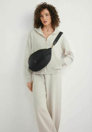 Kobieta o kręconych włosach, ubrana w kremową bluzę z zamkiem i spodnie, niosąca dużą czarną torbę na ramię na tle białego tła.