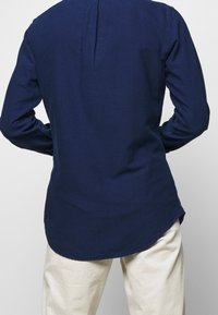 Navyblå långärmad skjorta i lätt tyg, med rundad avslutning och bakfåll, samt figursydda ärmar. Matchad med krämfärgade byxor.