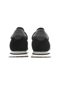 Adidași din suede negru au un tab de călcâi din piele netedă, cu inscripția „DIN 1890” debossată. Talpa intermediară albă are dungi negre pe margine.