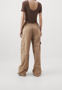 Braun sitzender Body mit tiefem Rückenausschnitt, kombiniert mit beigen Cargo-Hosen, die über Seitentaschen und einen entspannten Schnitt verfügen. Glattes Material, leichtes Textur.